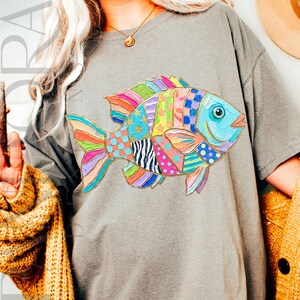 May include: A gray t-shirt featuring a colorful fish design. The fish is composed of various patterns and colors, including blue, pink, yellow, and green. The fish has a blue eye and a gold outline.