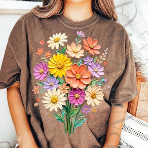 May include: Brown t-shirt featuring a vibrant floral bouquet design. The bouquet includes colorful flowers in shades of yellow, orange, pink, purple, and white. The flowers have a textured, 3D appearance, and the shirt has a vintage, worn look.