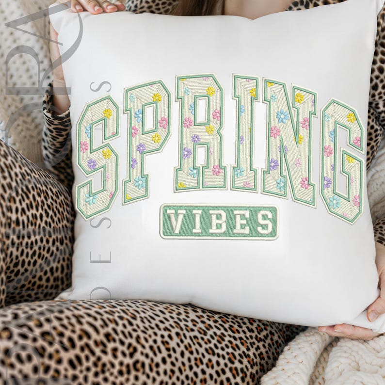 May include: White decorative pillow with the words "SPRING VIBES" embroidered in a green, floral design. The word "SPRING" is arched across the top, and "VIBES" is in a rectangular box below.