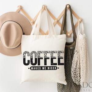 May include: A white tote bag with black text that reads "Coffee Makes Me Nicer." The text is in a leopard print font. The bag is hanging on a wooden rack with a brown hat and a beige mesh bag.