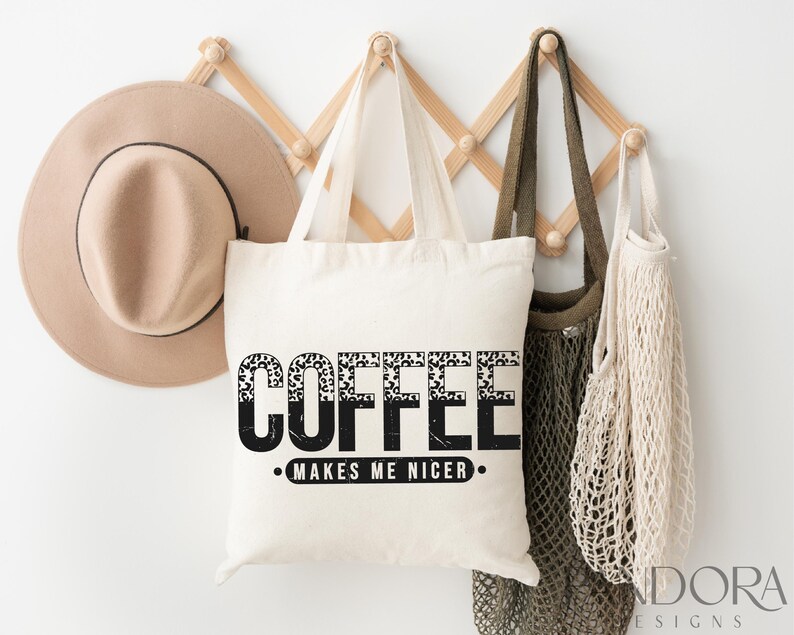 May include: A white tote bag with black text that reads "Coffee Makes Me Nicer." The text is in a leopard print font. The bag is hanging on a wooden rack with a brown hat and a beige mesh bag.