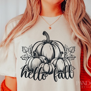 Peut inclure: Un t-shirt blanc avec un motif graphique noir de trois citrouilles et de feuilles. Le texte "hello fall" est &eacute;crit sous les citrouilles.
