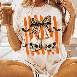 May include: A cream-colored t-shirt featuring a graphic of two human skulls hanging from a leopard-print bow. The design includes vertical orange brushstrokes, small black bats, and gold star accents. The skulls are positioned side by side.