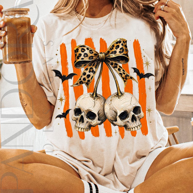 May include: A cream-colored t-shirt featuring a graphic of two human skulls hanging from a leopard-print bow. The design includes vertical orange brushstrokes, small black bats, and gold star accents. The skulls are positioned side by side.