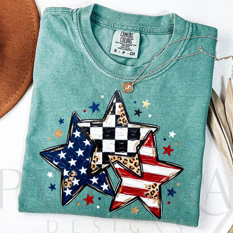 May include: A teal Comfort Colors t-shirt featuring three star designs with patriotic and leopard print patterns. The stars are outlined in black and have a gold border. A gold necklace with a small pendant is on the shirt. The shirt is folded on a white surface.