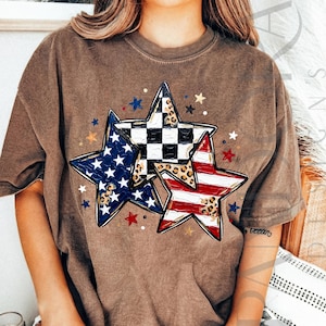 May include: Brown t-shirt featuring a graphic of three stars. One star has a blue and white pattern, another has a black and white checkered pattern, and the third has red and white stripes. Small stars in red, blue, and gold surround the larger stars.
