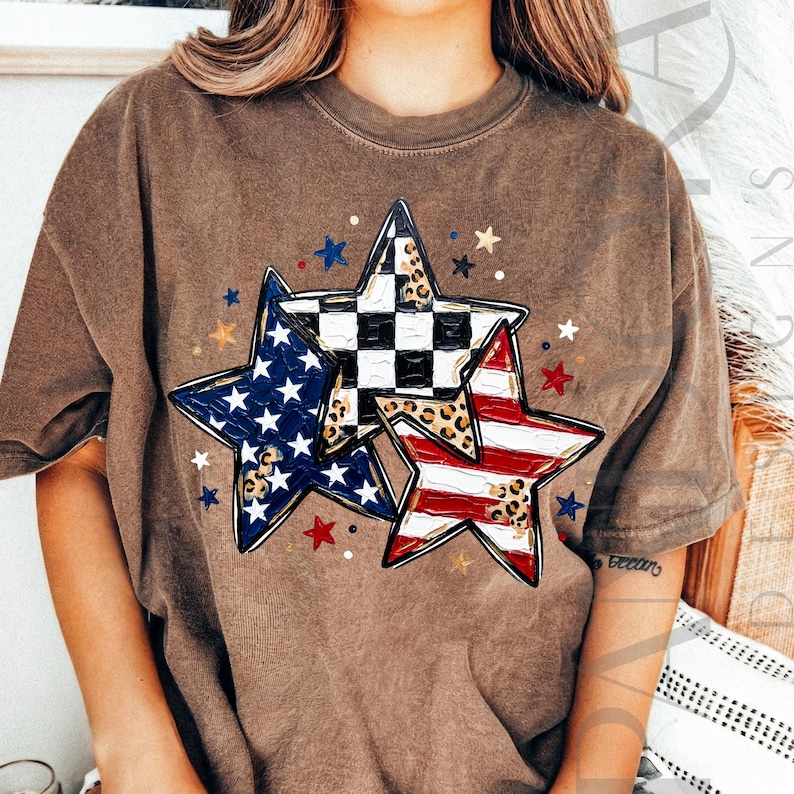 May include: Brown t-shirt featuring a graphic of three stars. One star has a blue and white pattern, another has a black and white checkered pattern, and the third has red and white stripes. Small stars in red, blue, and gold surround the larger stars.