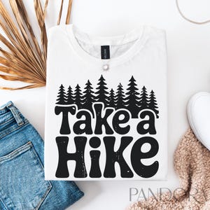 Może przedstawiać: Biała koszulka z czarnym nadrukiem przedstawiającym rząd drzew i tekst "Take a Hike" w retro czcionce.