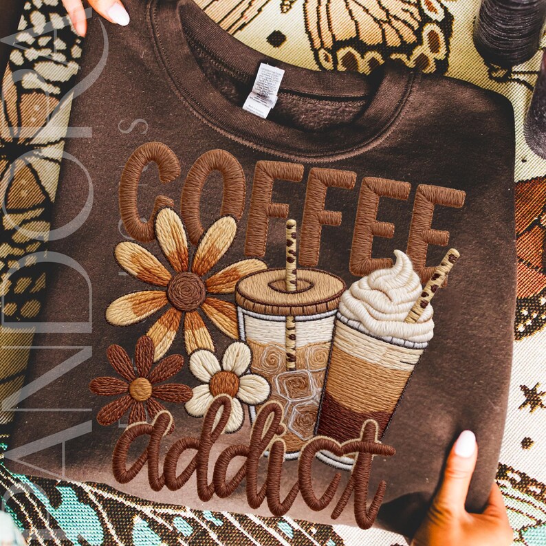 May include: A brown sweatshirt with embroidered text and graphics. The word "COFFEE" is stitched across the top, with "addict" below. Floral designs and coffee cup illustrations are also embroidered on the front.