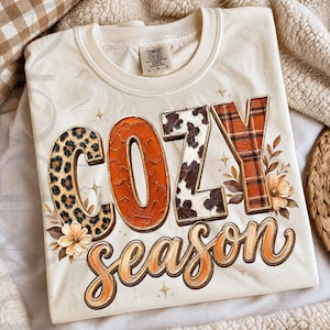 May include: A cream-colored t-shirt featuring the text COZY SEASON in large, stylized letters. The word COZY is printed with varied patterns including leopard print, textured orange, cow print, and orange plaid, accented by small floral illustrations.