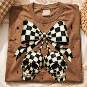 May include: A brown t-shirt featuring a graphic print of two skulls connected by stems to a large bow. Both the skulls and the bow have a black and white checkered pattern with gold outlines. Small four-pointed star icons surround the design.