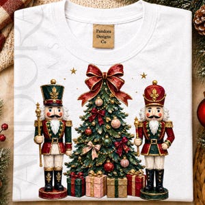 May include: A white t-shirt featuring a graphic of a decorated Christmas tree flanked by two nutcracker figures. The tree is topped with a large red bow and surrounded by wrapped gifts. The neck label reads Pandora Designs Co.