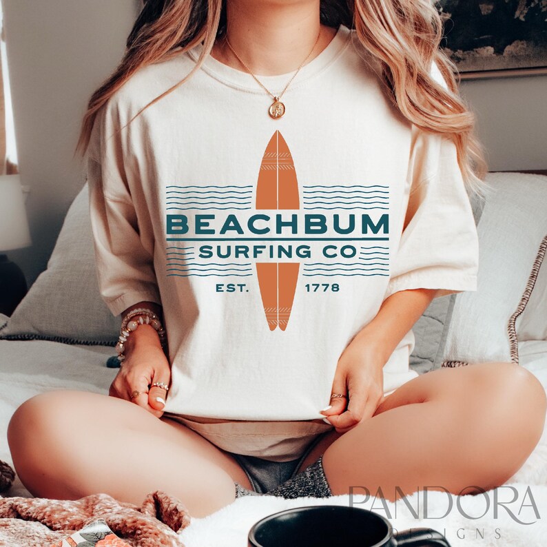 May include: Cream-colored t-shirt with a surfboard graphic and the text "BEACHBUM SURFING CO." in teal. The graphic includes a brown surfboard design and the year "1778". The shirt is a relaxed fit.