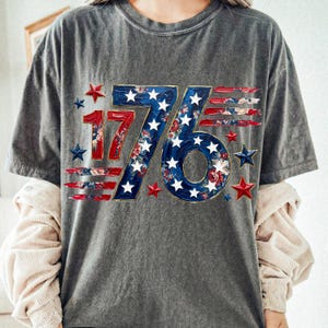 May include: Gray t-shirt with a patriotic "1776" design. The numbers are red and blue, with a floral pattern and white stars. Red and blue stars surround the design, creating a festive look.