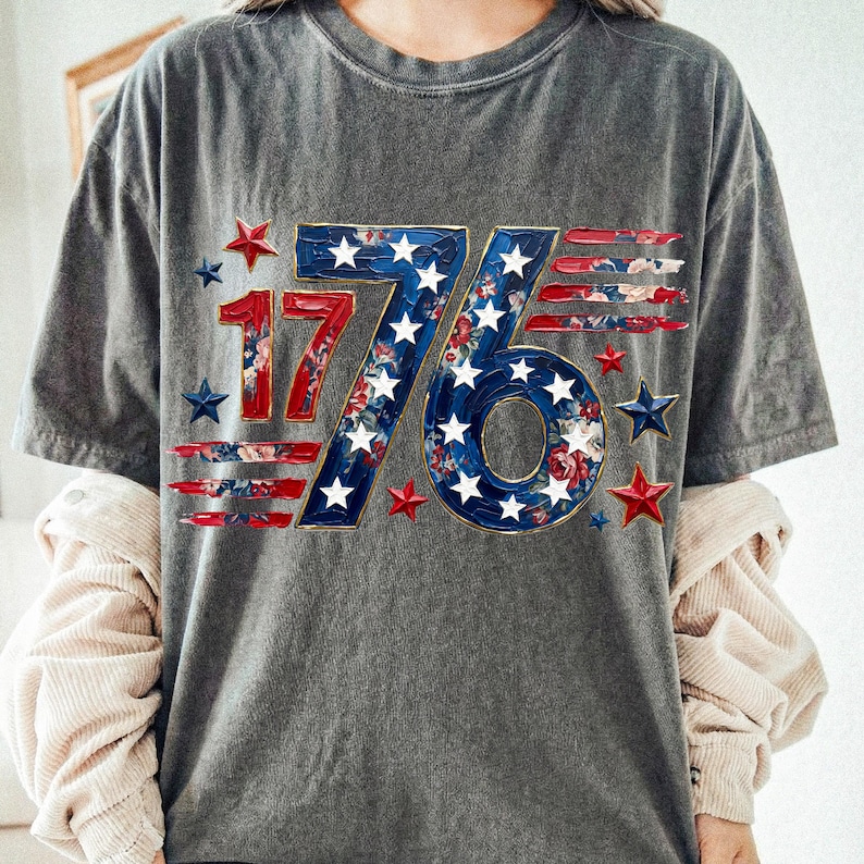 May include: Gray t-shirt with a patriotic "1776" design. The numbers are red and blue, with a floral pattern and white stars. Red and blue stars surround the design, creating a festive look.