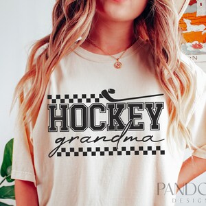 May include: A cream-colored t-shirt with black text that reads "HOCKEY grandma" with a black and white checkered pattern above and below the text. A black hockey stick with a puck is above the text.