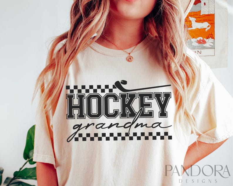 May include: A cream-colored t-shirt with black text that reads "HOCKEY grandma" with a black and white checkered pattern above and below the text. A black hockey stick with a puck is above the text.
