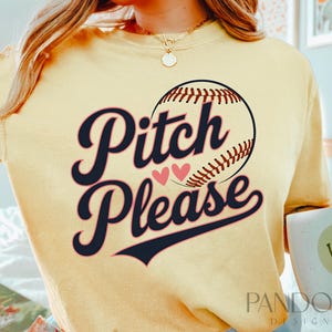 K&ouml;nnte beinhalten: Ein gelbes T-Shirt mit einem Baseball-Motiv und dem Text "Pitch Please" in Schwarz-Wei&szlig;. Zwei rosa Herzen befinden sich &uuml;ber dem Text.