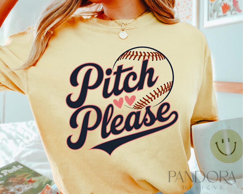 K&ouml;nnte beinhalten: Ein gelbes T-Shirt mit einem Baseball-Motiv und dem Text "Pitch Please" in Schwarz-Wei&szlig;. Zwei rosa Herzen befinden sich &uuml;ber dem Text.