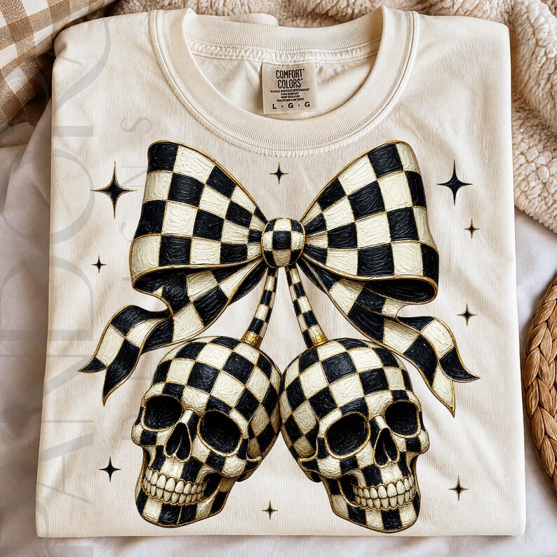 May include: A cream-colored t-shirt featuring a graphic of two skulls and a large bow, all decorated with a black and white checkered pattern. The design includes small four-pointed star accents. The shirt has a Comfort Colors brand label.