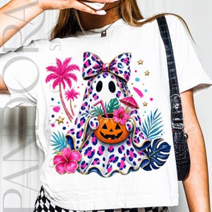May include: A white t-shirt featuring a graphic of a ghost covered in a pink and blue leopard print pattern. The ghost wears a matching bow, holds an orange jack-o'-lantern drink with a straw and umbrella, and is surrounded by palm trees and hibiscus flowers.