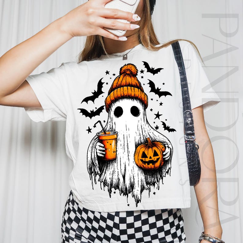May include: White t-shirt with a Halloween ghost design. The ghost wears an orange beanie and holds a pumpkin and a drink. Black bats and stars are also featured. The ghost has black eyes and a white sheet-like body.