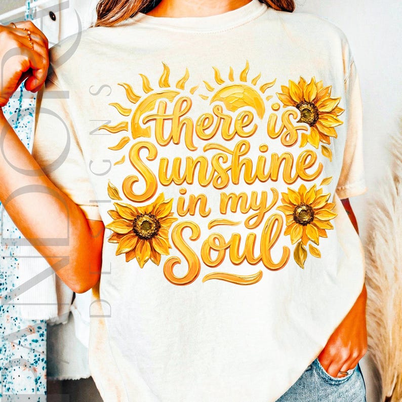 May include: Cream-colored t-shirt with a vibrant graphic featuring sunflowers and the phrase "there is Sunshine in my Soul" in a decorative font. The design includes two suns and three sunflowers in shades of yellow and orange.