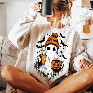 May include: Cream sweatshirt with a Halloween ghost design. The ghost wears an orange beanie and holds a pumpkin and a drink. Black bats and stars surround the ghost. The sweatshirt is a cosy, festive garment for the autumn season.
