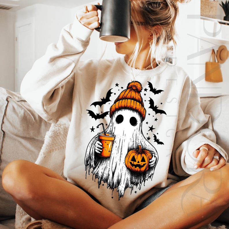 May include: Cream sweatshirt with a Halloween ghost design. The ghost wears an orange beanie and holds a pumpkin and a drink. Black bats and stars surround the ghost. The sweatshirt is a cosy, festive garment for the autumn season.