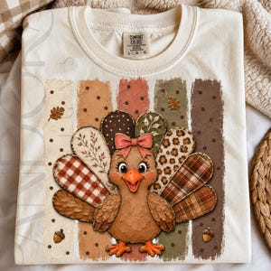 May include: A cream-colored t-shirt featuring a central graphic of a brown turkey with a pink bow. The turkey has tail feathers in various patterns including plaid, leopard print, and floral. Vertical textured stripes and small acorns surround it.