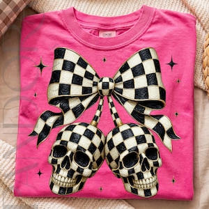 May include: A bright pink t-shirt featuring a large graphic of two skulls connected by stems to a central bow. The skulls and bow are decorated with a black and white checkered pattern. Small black four-pointed star accents surround the design.