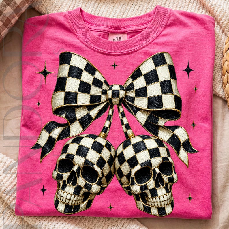 May include: A bright pink t-shirt featuring a large graphic of two skulls connected by stems to a central bow. The skulls and bow are decorated with a black and white checkered pattern. Small black four-pointed star accents surround the design.
