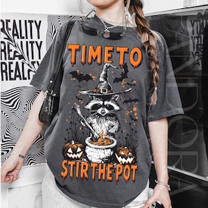 May include: Dark gray t-shirt with a Halloween-themed graphic. The design features a raccoon wearing a witch hat, stirring a cauldron, with the text "TIME TO STIR THE POT" in orange. Bats and pumpkins complete the spooky design.