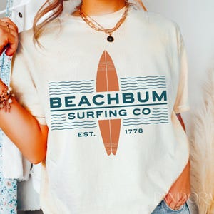 May include: Cream-colored t-shirt with a graphic design featuring a surfboard and the text "BEACHBUM SURFING CO." in teal. The design includes the year "1778" and the word "EST." The shirt has a relaxed fit.