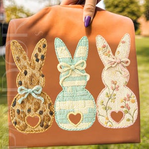 May include: A brown fabric item featuring three embroidered bunny silhouettes. Each bunny has a heart-shaped cutout and a bow. The bunnies are in shades of brown, blue, and pink, with floral and patterned details. The background is a solid orange color.