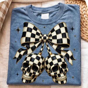 May include: A blue t-shirt featuring a graphic print of two skulls and a large bow, all decorated with a black and white checkered pattern. The design includes small four-pointed star accents scattered around the bow and skulls.