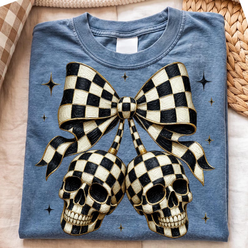 May include: A blue t-shirt featuring a graphic print of two skulls and a large bow, all decorated with a black and white checkered pattern. The design includes small four-pointed star accents scattered around the bow and skulls.