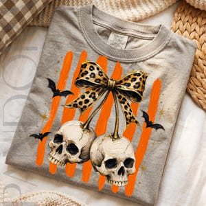 May include: A grey t-shirt featuring a graphic of two off-white skulls hanging from a leopard print bow. The design includes vertical orange brushstrokes, small black bats, and gold star accents. The inner neck label reads Comfort Colors.