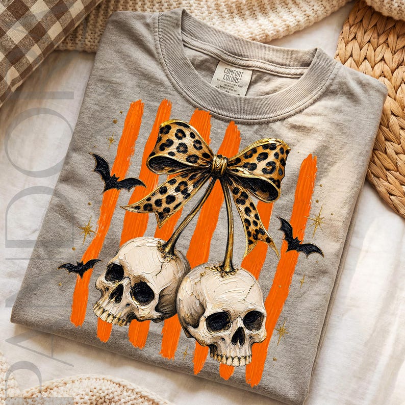 May include: A grey t-shirt featuring a graphic of two off-white skulls hanging from a leopard print bow. The design includes vertical orange brushstrokes, small black bats, and gold star accents. The inner neck label reads Comfort Colors.