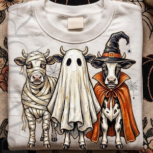 Può includere: Felpa bianca con un design a tema Halloween di tre mucche in costume. Una è una mummia, un'altra un fantasma e la terza un vampiro con mantello e cappello da strega. L'illustrazione ha uno stile cartoon.