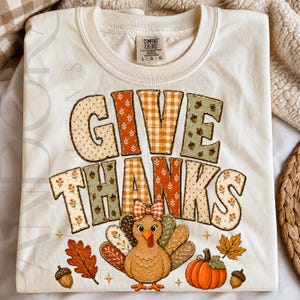 Pode incluir: Uma t-shirt em tom creme com o texto GIVE THANKS em letras estampadas com motivos de outono. Abaixo do texto, há um peru de desenho animado com um laço xadrez, acompanhado por uma abóbora laranja, bolotas e folhas.