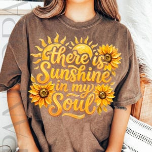 May include: Brown t-shirt with a graphic design featuring the text "There is Sunshine in my Soul" in yellow script, along with yellow sunflowers and sun illustrations. The shirt has a vintage, worn look.