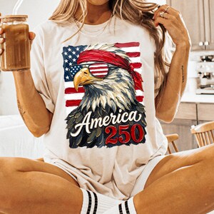 Puede incluir: Camiseta color crema con un gr&aacute;fico de un &aacute;guila calva americana con gafas de sol con dise&ntilde;o de rayas rojas y blancas. El &aacute;guila est&aacute; sobre la bandera americana, con el texto "America 250" debajo.