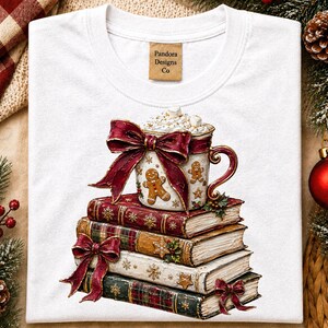 Op de afbeelding: Een wit t-shirt met een afbeelding van een stapel van drie boeken met daarop een mok warme chocolademelk. De mok is versierd met een peperkoekmannetje en een grote rode strik. De boeken zijn voorzien van gouden sneeuwvlokken en ruitpatronen.