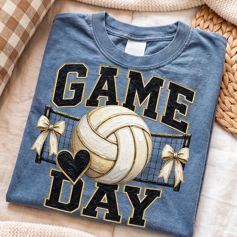 May include: A blue crew neck t-shirt featuring the text GAME DAY in black block letters with gold outlines. Centered is a white volleyball graphic, a black heart, a black volleyball net, and two cream-colored bows on either side of the net.