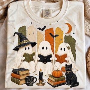 May include: A cream-colored t-shirt featuring a graphic of three white ghosts reading books. One ghost wears a black witch hat. Below are stacks of books, a black skull mug, a black candle in a holder, an orange pumpkin, and a black cat.