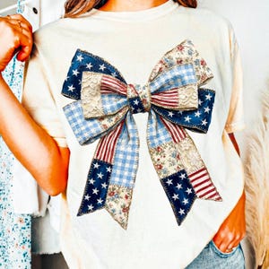 Pu&ograve; includere: T-shirt color crema con un grande motivo a fiocco. Il fiocco &egrave; un patchwork di motivi patriottici, tra cui blu con stelle bianche, strisce rosse e bianche e stampe floreali. Il centro del fiocco ha un dettaglio in pizzo.