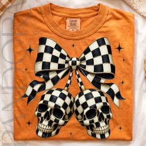 May include: An orange t-shirt featuring a graphic of two skulls and a large bow, all decorated with a black and white checkered pattern. The design includes small black four-pointed stars scattered around the central skull and bow illustration.