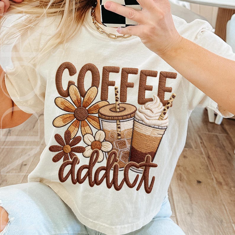 This design features the words 'Coffee Addict' in bold, retro-style lettering in warm brown tones. The graphic features two iced coffee cups complete with straws and whipped cream, surrounded by large floral elements in neutral brown shades. The overall aesthetic is trendy yet cosy, drawing inspiration from boutique-style apparel for coffee lovers.