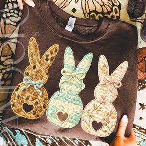 May include: A brown sweatshirt featuring three embroidered bunny designs. Each bunny has a heart-shaped cutout and a bow. The bunnies are in shades of tan, mint green, and pink, with floral and patterned details. The sweatshirt is a cozy, casual garment.